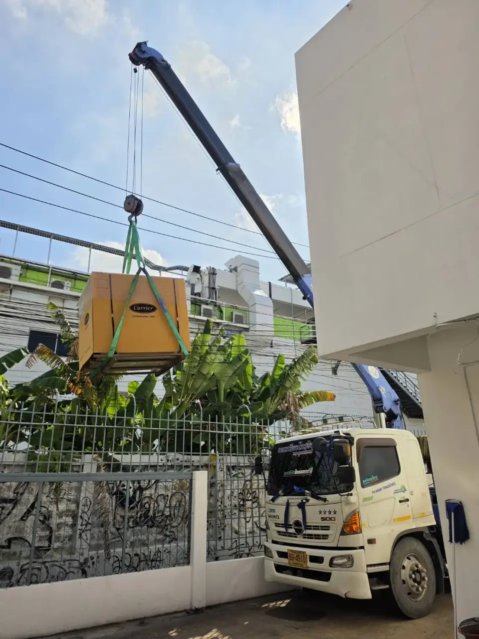 รถเฮี๊ยบ 5 ตัน ยกเครื่องปรับอากาศด้วยสลิงผ้าบริเวณข้างอาคารและรั้วเหล็ก